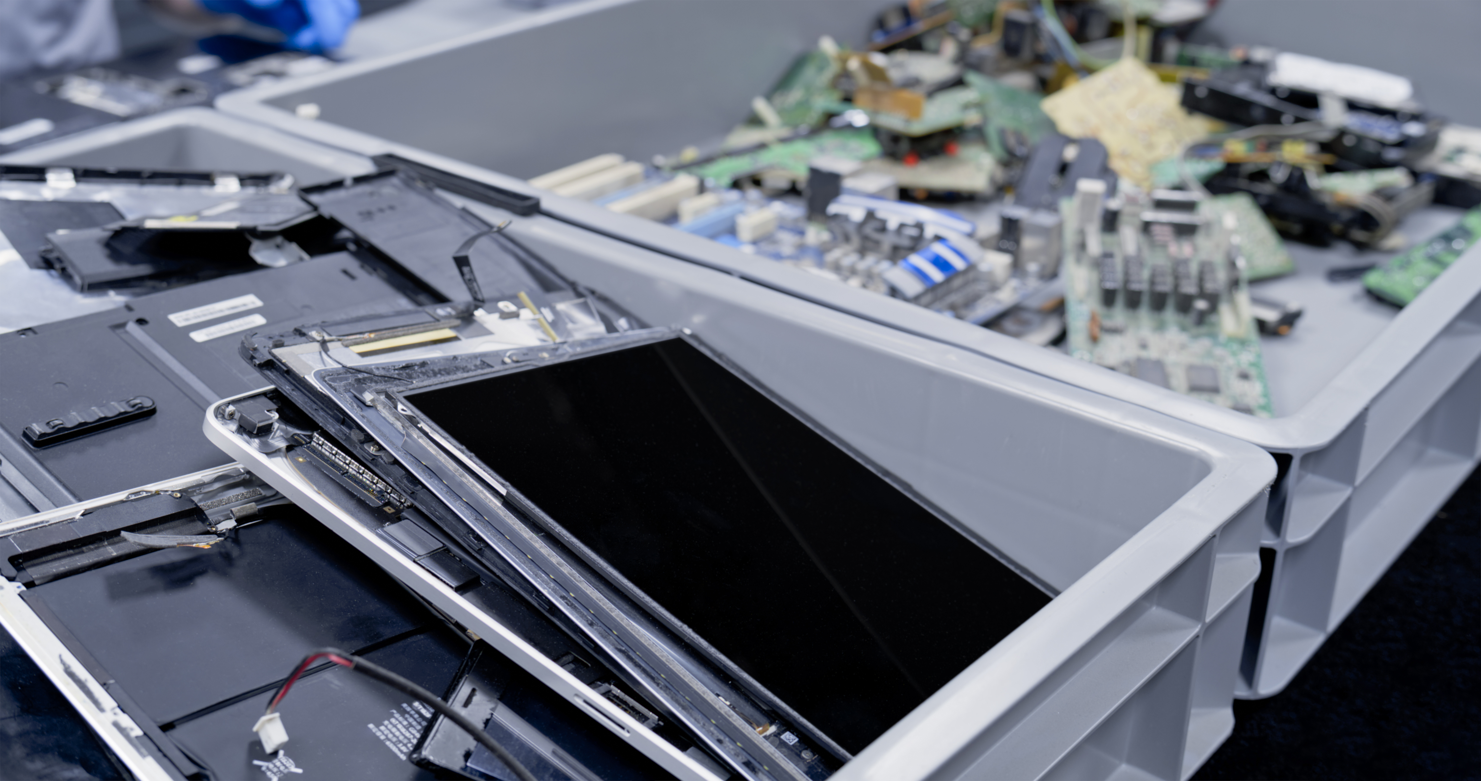 Disassembled electronic devices in gray plastic bins, including tablets and circuit boards, in a recycling or repair facility.
